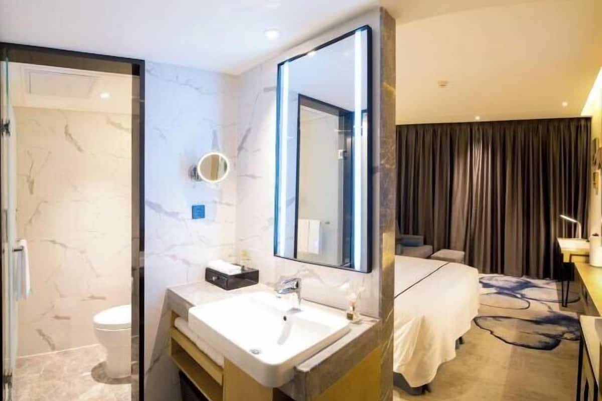 A modern hotel room with a bathroom, a large mirror, a bed, and a desk.