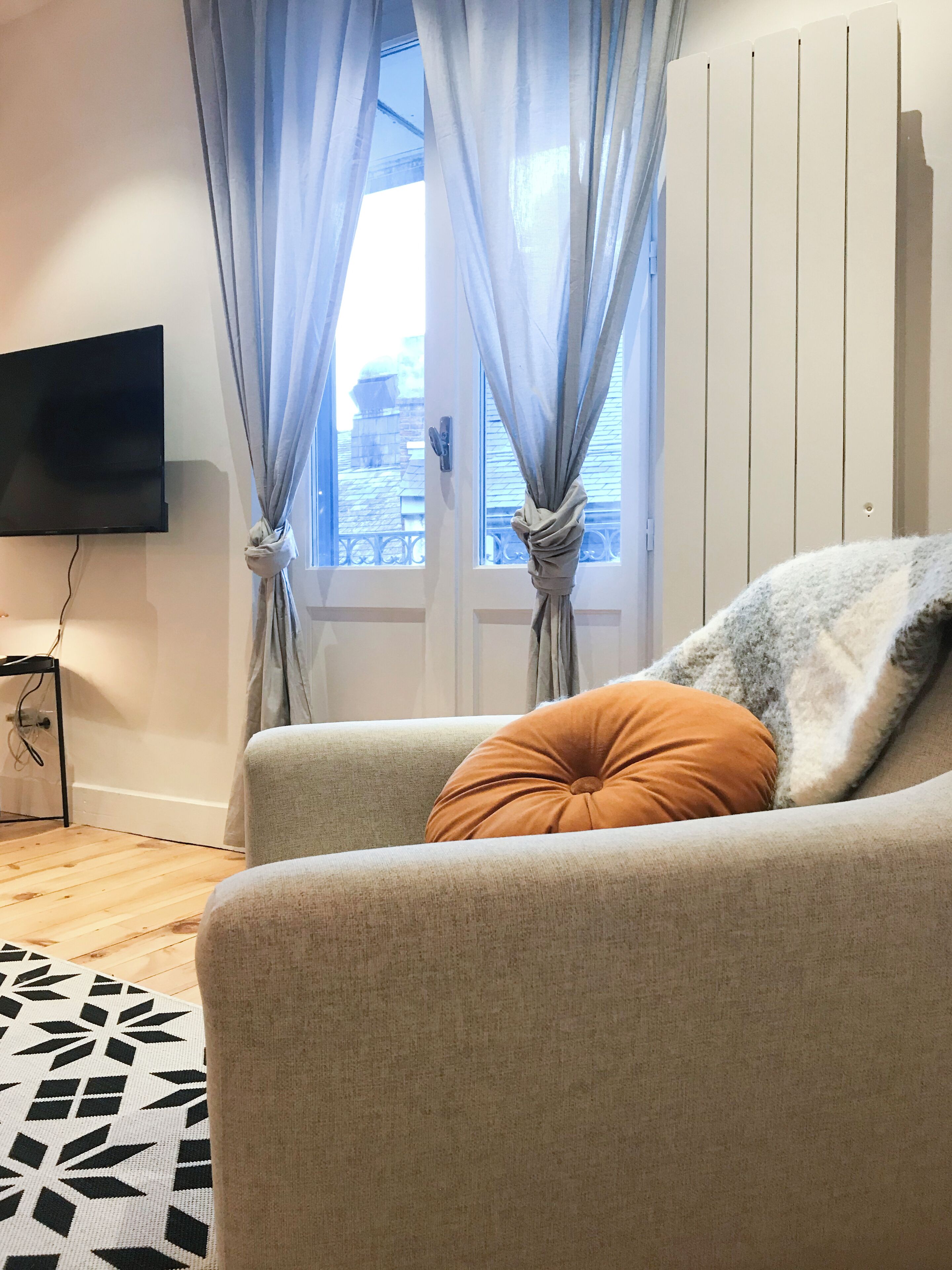 Un salon avec un canapé, un téléviseur fixé au mur et une fenêtre avec des rideaux.