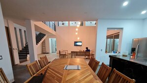 A spacious dining area with a large wooden table and chairs, a staircase in the background, and a kitchen area with a refrigerator and sink.