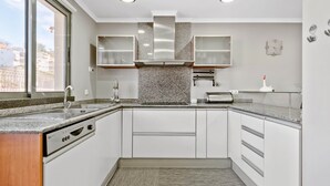 A modern kitchen with a dishwasher, granite countertops, white cabinetry, a stainless steel range hood, and a window providing a view of the outside. - Penthouse Prestige by Homie (Funchal)