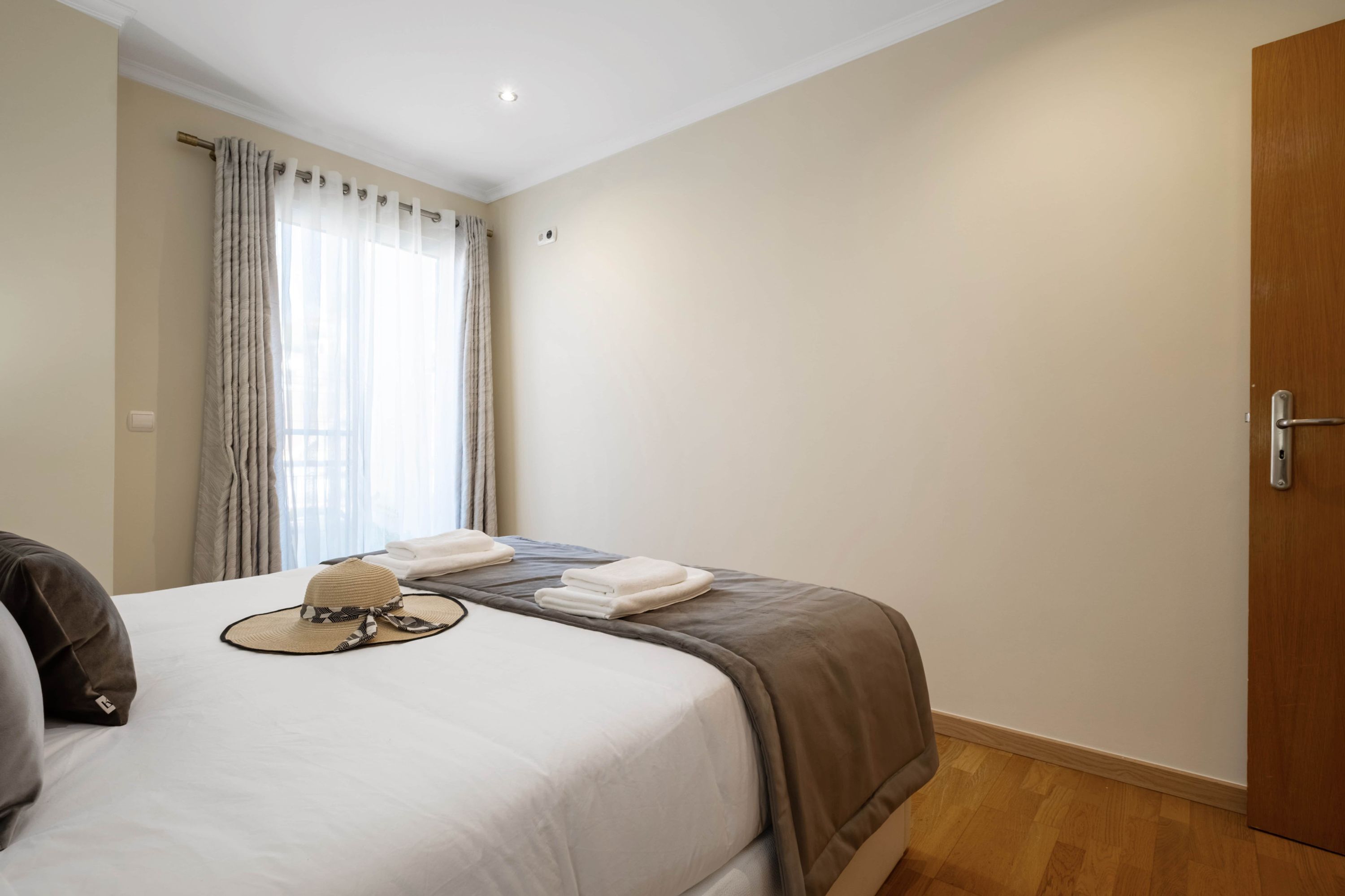 A hotel room with a bed, a window with curtains, a door, and a wooden floor.