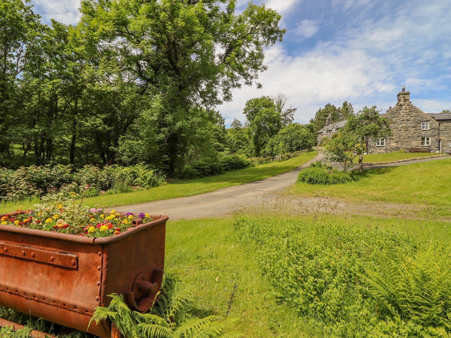 A rural road leading to a stone house with a stone chimney, surrounded by lush greenery and a flower bed.