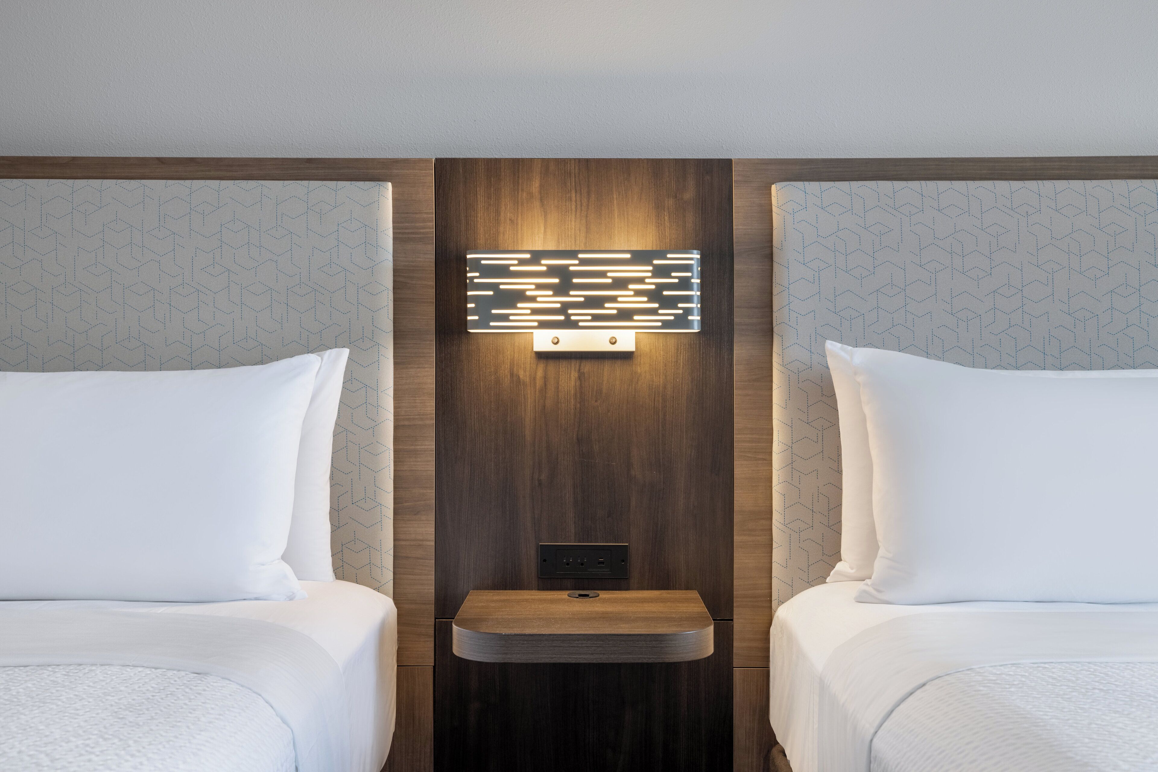 A hotel room with two beds, a wall-mounted light panel, and a small wooden shelf.