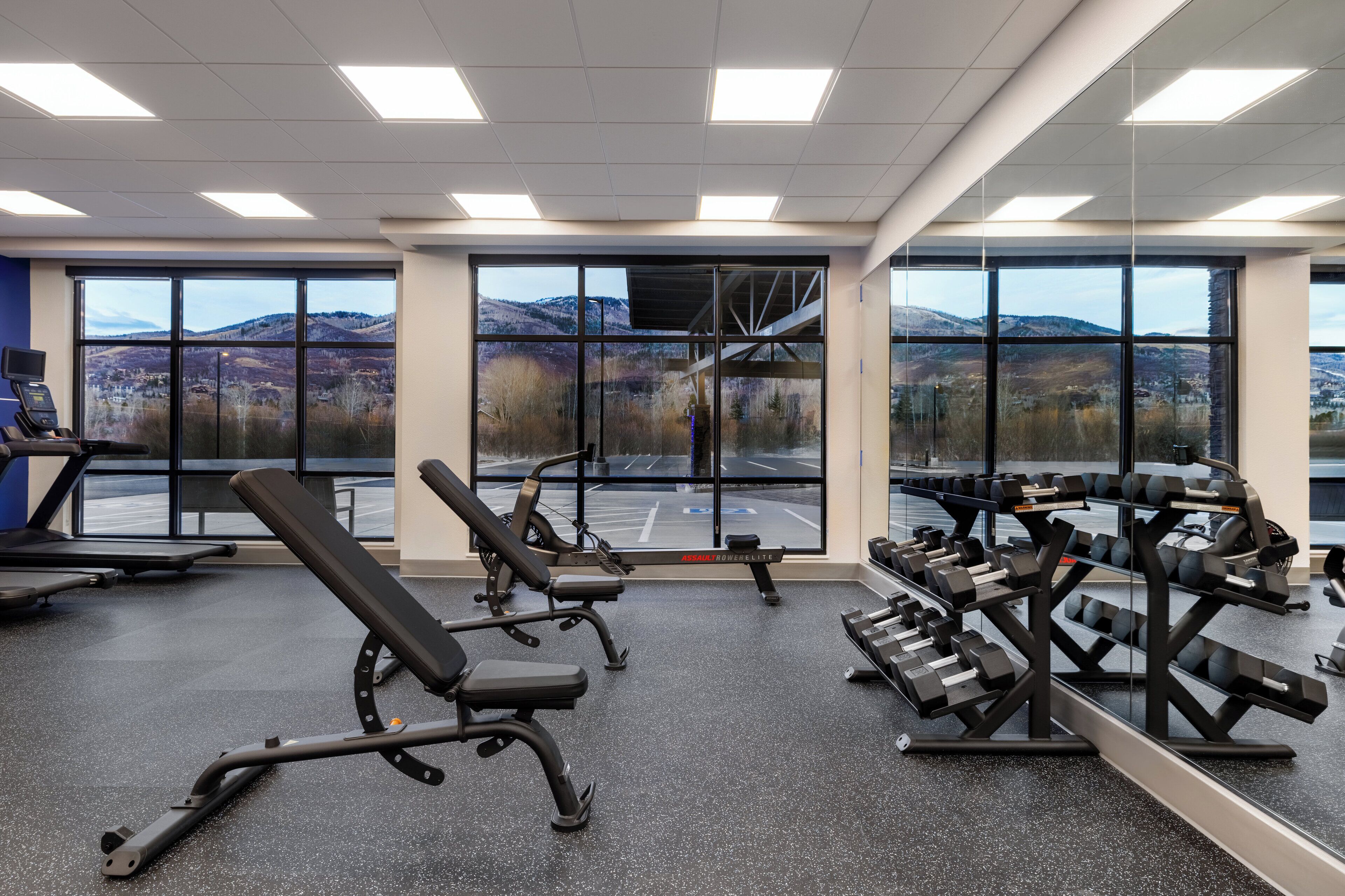 Un gimnasio con equipo de ejercicio, ventanas amplias y vistas a montañas.