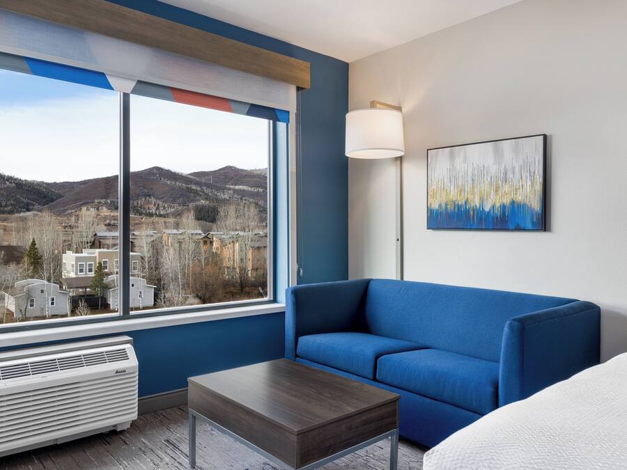 A hotel room with a large window, a blue sofa, a bed, a bedside table, a lamp, and a painting.
