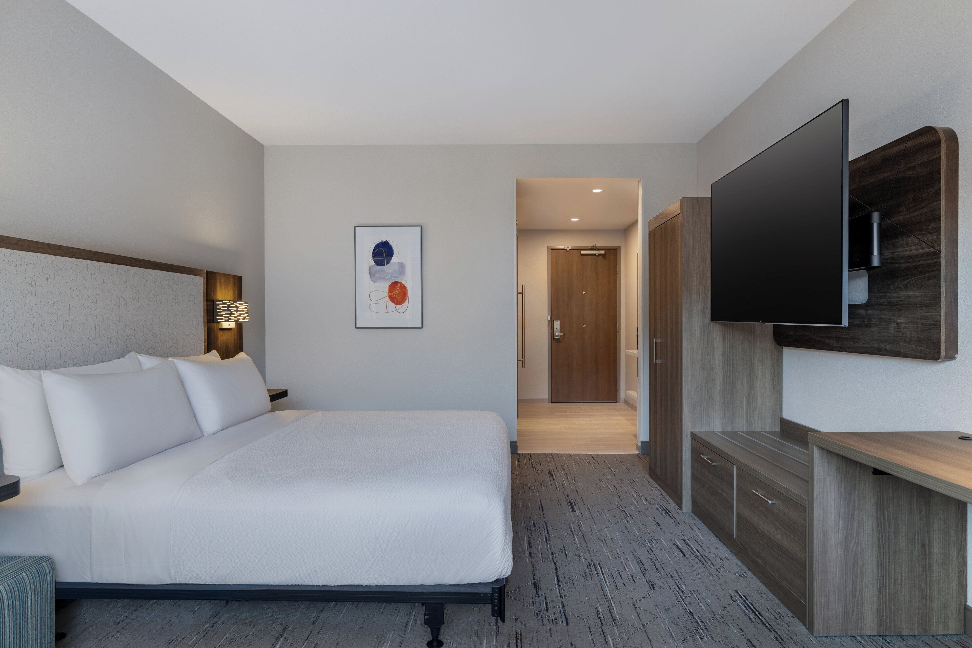 A modern hotel room with a large bed, a flat-screen TV mounted on the wall, a wooden cabinet, and a framed artwork on the wall.
