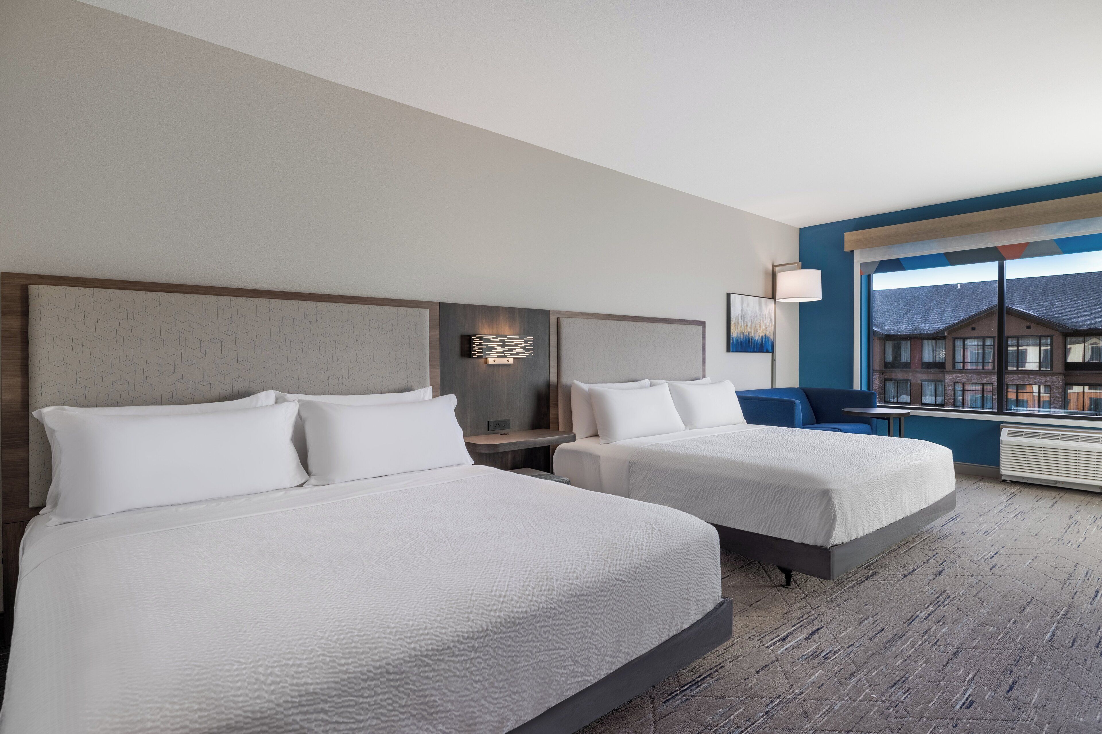 A hotel room with two beds, a desk, a chair, a lamp, and a window with a view of buildings.