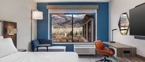 A hotel room with a large window offering a mountain view, a bed, a desk, and a chair.