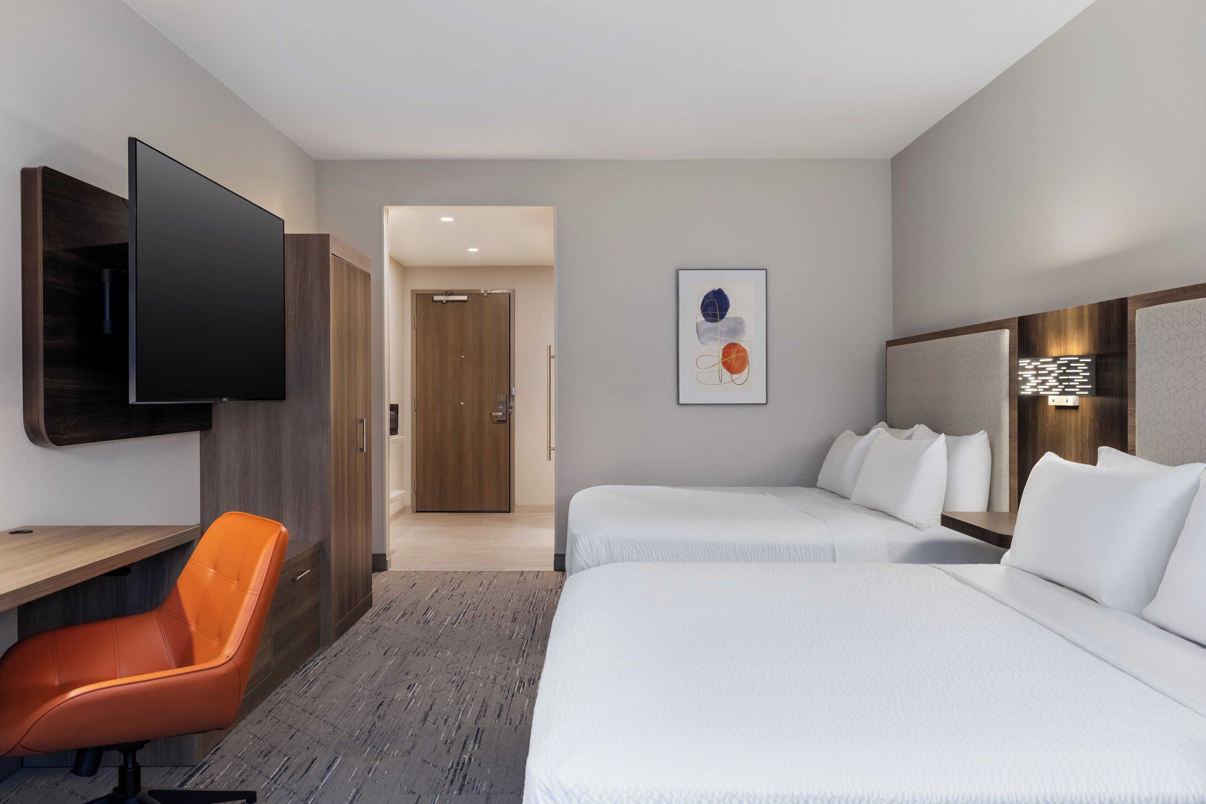 Ein Hotelzimmer mit zwei Betten, einem Schreibtisch, einem Stuhl, einem Flachbildfernseher und einer Tür zu einem weiteren Raum.