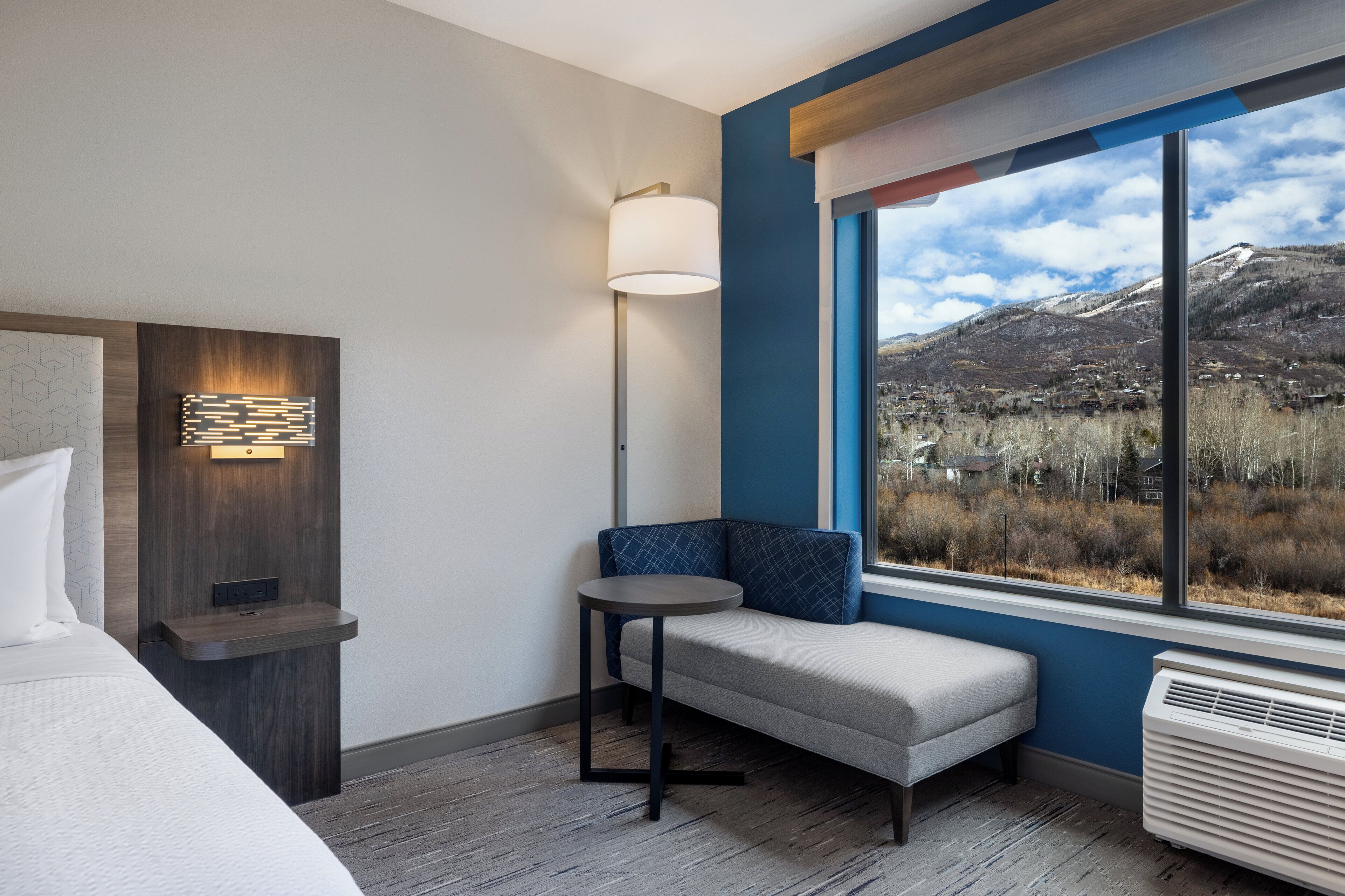A hotel room with a bed, a small table, a bench, a wall-mounted lamp, and a large window offering a mountain view.