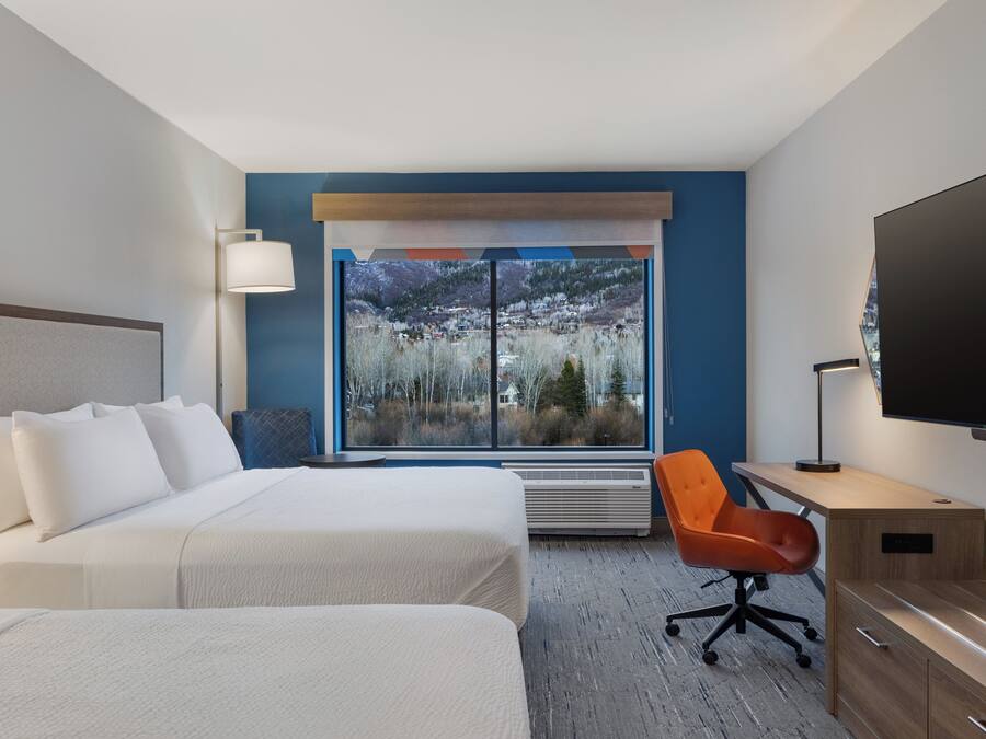 A hotel room with two beds, a desk with a chair, a TV, and a large window with a view of a snowy landscape.