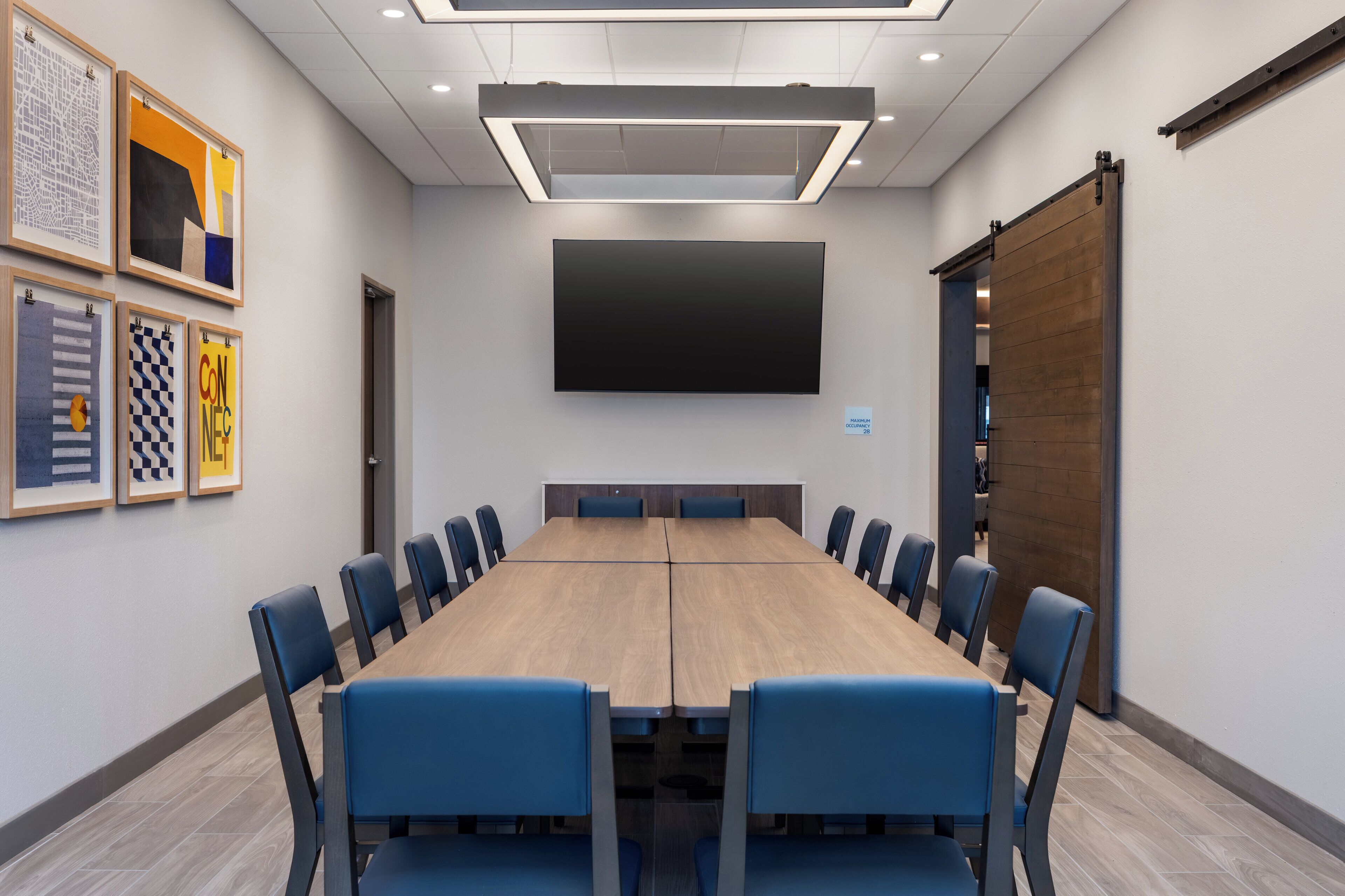A conference room with a long table, blue chairs, a large flat-screen TV, and framed artwork on the walls.