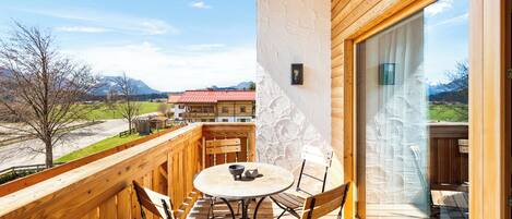Un balcón de madera con una mesa redonda y sillas, vista a una montaña y un edificio con techo rojo al fondo.