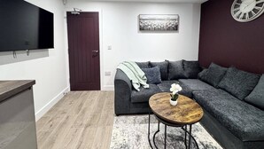 Ein modernes Wohnzimmer mit einem grauen Ecksofa, einem Couchtisch aus Holz mit einer Pflanze, einem wandmontierten Flachbildfernseher, einem gerahmten Bild an der Wand und einer Uhr.