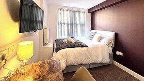 Ein Hotelzimmer mit einem Bett, einem an der Wand montierten Fernseher, einem Fenster mit Jalousien, einer Lampe und einem Nachttisch aus Holz.