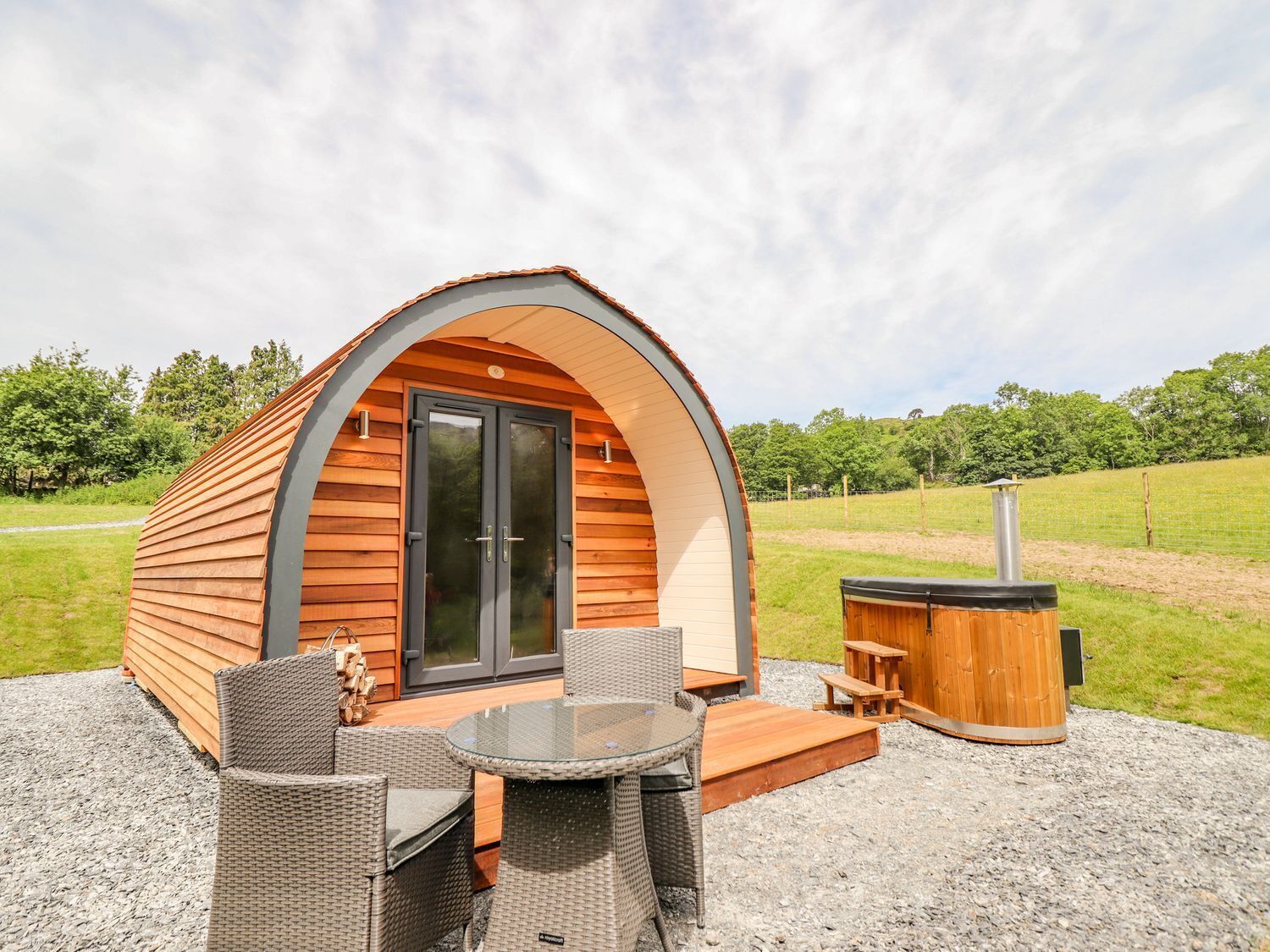A cozy, semi-open wooden cabin with a glass door, furnished with wicker chairs and a glass table, set on a gravel surface with a barbecue grill and a hot tub nearby.
