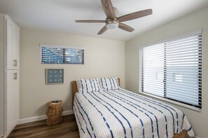Une chambre à coucher avec un lit, un ventilateur de plafond, une fenêtre avec des stores, un panier en osier et un tableau encadré au mur.