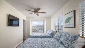 Un dormitorio con ventilador de techo, cama con ropa de cama de diseño azul y blanco, televisor montado y un cuadro de una tortuga enmarcada.