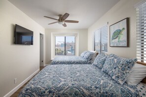 Ein Schlafzimmer mit Deckenventilator, einem Bett mit blau-weiß gestreifter Bettwäsche, einem an der Wand befestigten Fernseher und einem gerahmten Gemälde von einer Schildkröte.
