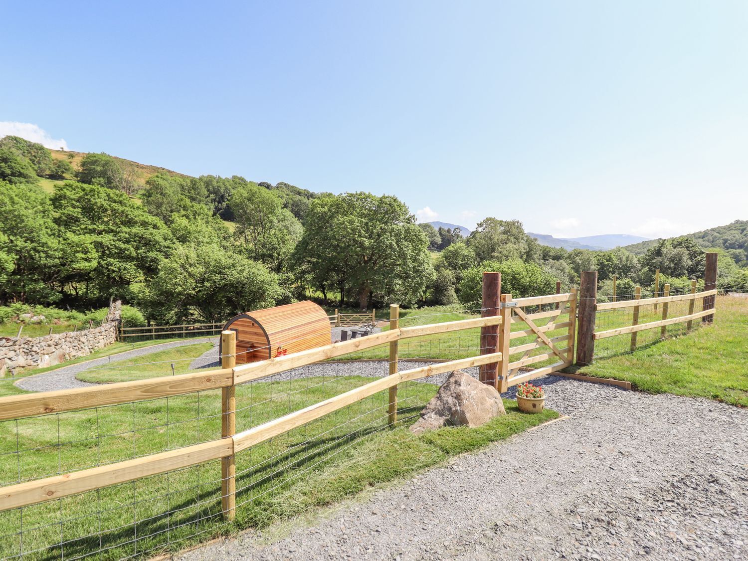 Un recinto in legno con una struttura coperta, probabilmente un'antica carrozza o un edificio per le carrozze, in un contesto rurale con dolci colline e alberi sullo sfondo.