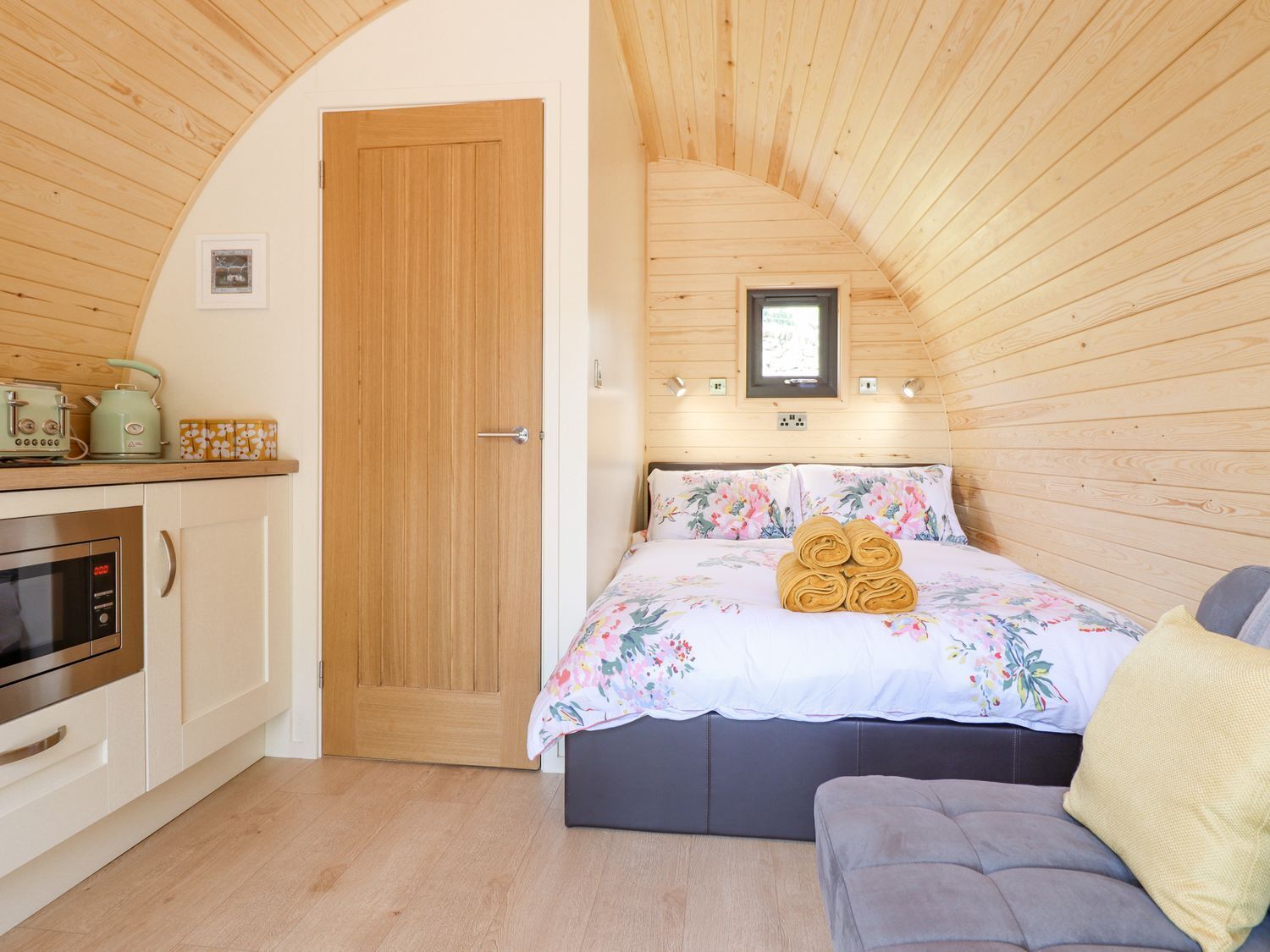 A cozy bedroom with a wooden ceiling, a bed with floral bedding, a bedside table, and a small window.