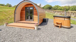 A small, single-story wooden cabin with a curved roof, a front door, and a patio area with a bench and a hot tub. - Eirlys (Dolgellau)