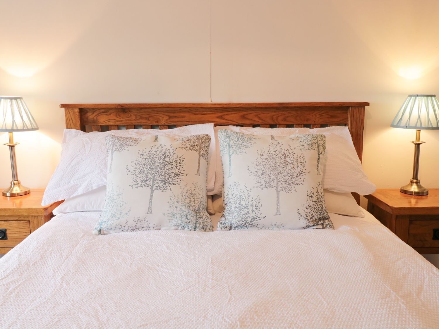 Een net opgemaakt bed met witte lakens en twee bijpassende kussens met een boommotief, geflankeerd door twee nachtkastjes met lampen.