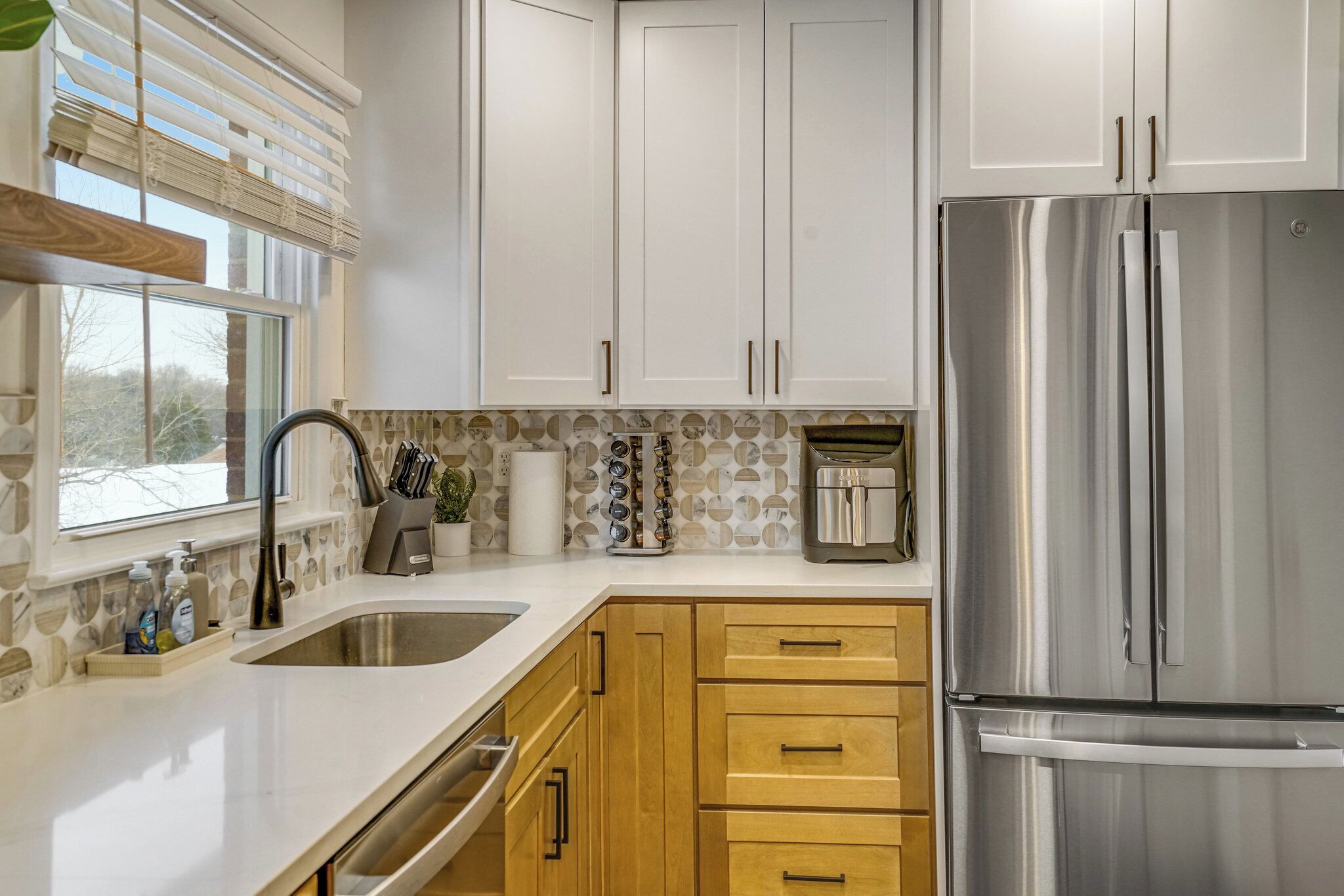 Una cocina con armarios blancos, un refrigerador de acero inoxidable, un lavamanos y una ventana con persianas.