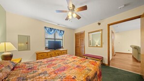Ein Schlafzimmer mit einem Bett, einem Deckenventilator, einem Fernseher auf einer Kommode, einem Fenster mit Vorhängen, einem Spiegel und einer Tür zu einem anderen Raum.