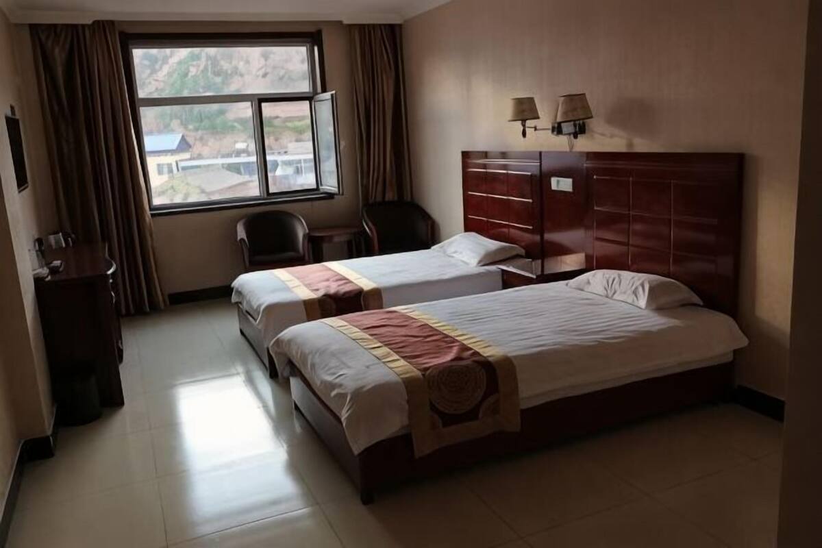 A hotel room with two beds, a desk, a chair, a window with curtains, and a lamp.