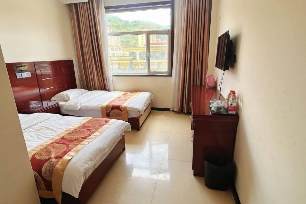 A hotel room with two beds, a TV, a desk with a trash bin, and a window with curtains.