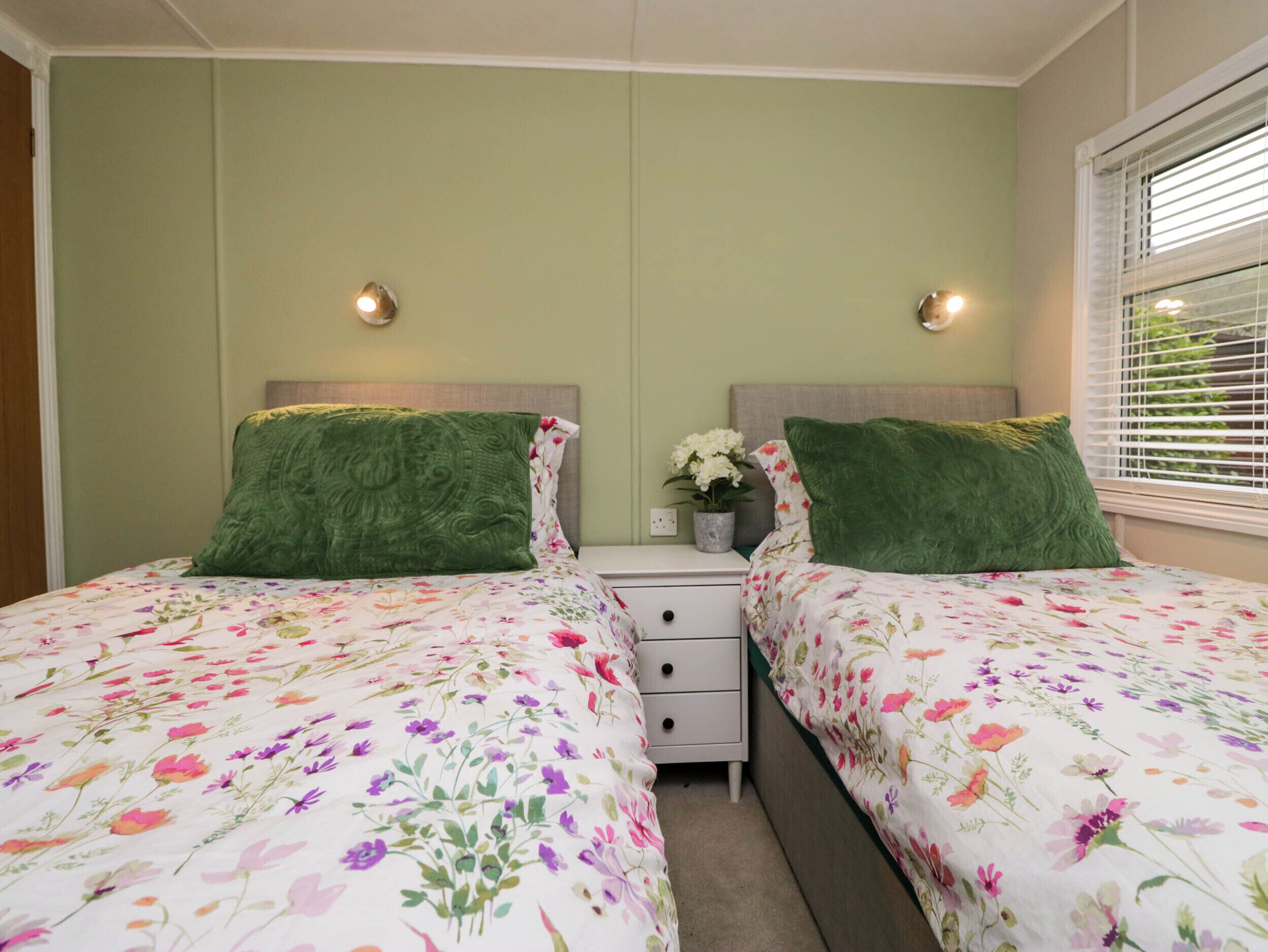 A twin bedroom with floral bedding, a nightstand with a vase of flowers, and two wall-mounted lights.