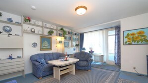 A living room with a blue sofa, a patterned rug, a white coffee table, and a bookshelf with various decorative items. - House Captain Christiansen 53 (Westerland)