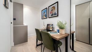 A modern dining area with a wooden table, two framed artworks, a laptop, a notebook, a pen, a potted plant, and a green chair. - Camden | Room B2 - Studio Apartment, Sleeps 2 (London)