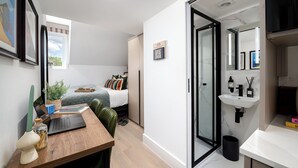 A modern, compact living space with a bed, a desk with a laptop, a small plant, and a minimalist bathroom with a sink and mirror. - Camden | Room B2 - Studio Apartment, Sleeps 2 (London)
