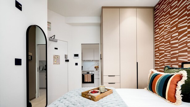 A modern bedroom with a large bed, a wardrobe, a mirror, and a kitchenette in the background. - Camden | Studio W Separate Kitchen (London)