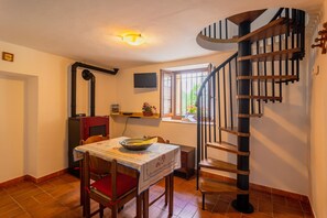Un coin repas avec une table dressée pour deux personnes, une chaise en bois, un coussin rouge, une petite table sur laquelle se trouvent un bol et un livre, une fenêtre donnant sur un paysage verdoyant et un escalier en colimaçon.