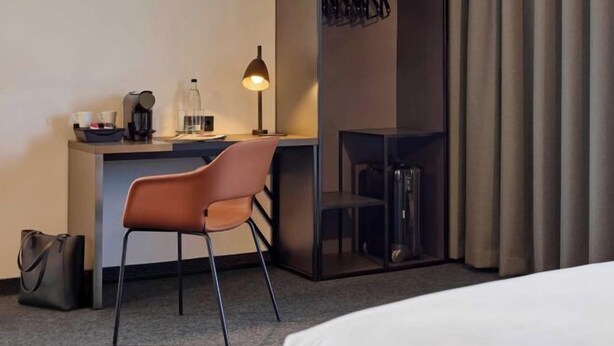 A hotel room with a bed, a desk with a chair, a lamp, a coat rack, and a suitcase. - B&B HOTEL München City-Nord (Munich)
