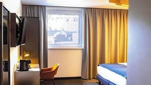 A hotel room with a bed, a desk with a lamp, a chair, and a window with curtains. - B&B HOTEL München City-Nord (Munich)