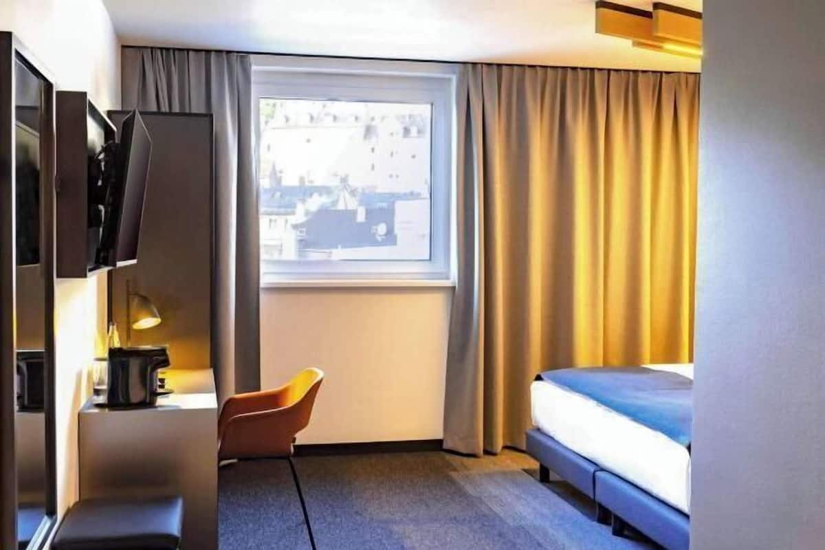 Una habitación de hotel con una cama, un escritorio con lámpara, una silla y una ventana con cortinas.