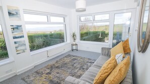 A room with large windows, a grey sofa with cushions, a small table with a plant, and a radiator. - Burry House (Swansea)