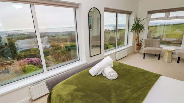 A bedroom with a large window, a bed with a green blanket, a chair, a small table, and a potted plant. - Burry House (Swansea)