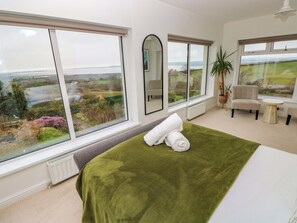 A bedroom with a large window, a bed with a green blanket, a chair, a small table, and a potted plant. - Burry House (Swansea)