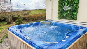 A hot tub in a backyard with a wooden deck, surrounded by a grassy area and a decorative wall with greenery. - Burry House (Swansea)