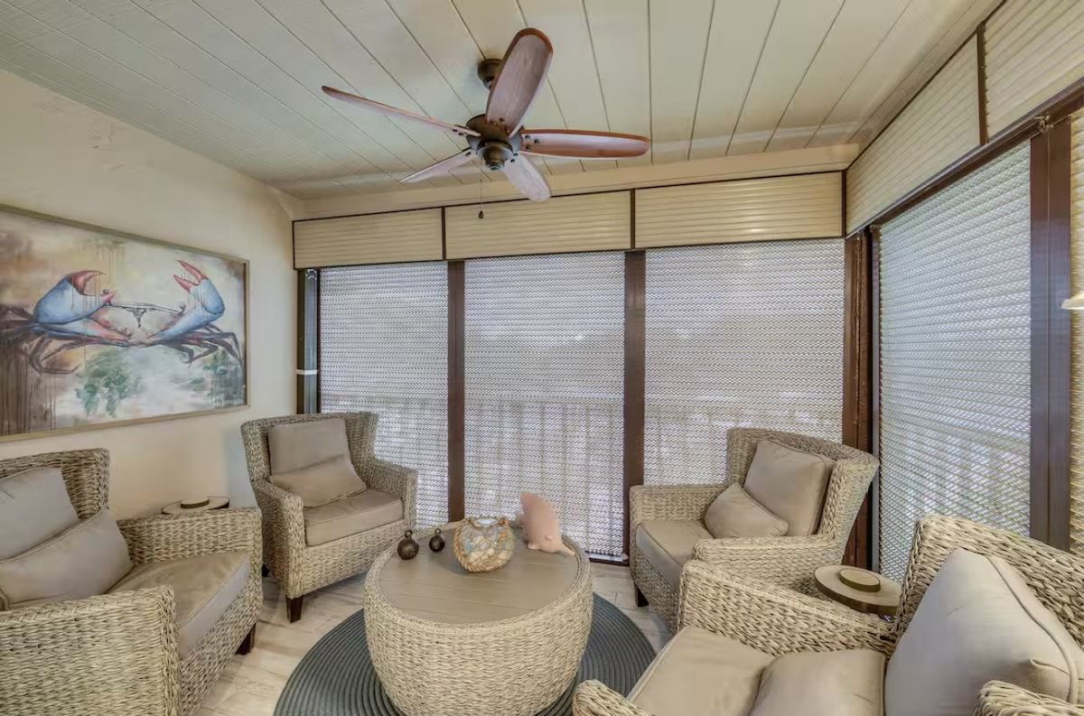 A room with wicker furniture, a ceiling fan, and large windows with blinds.
