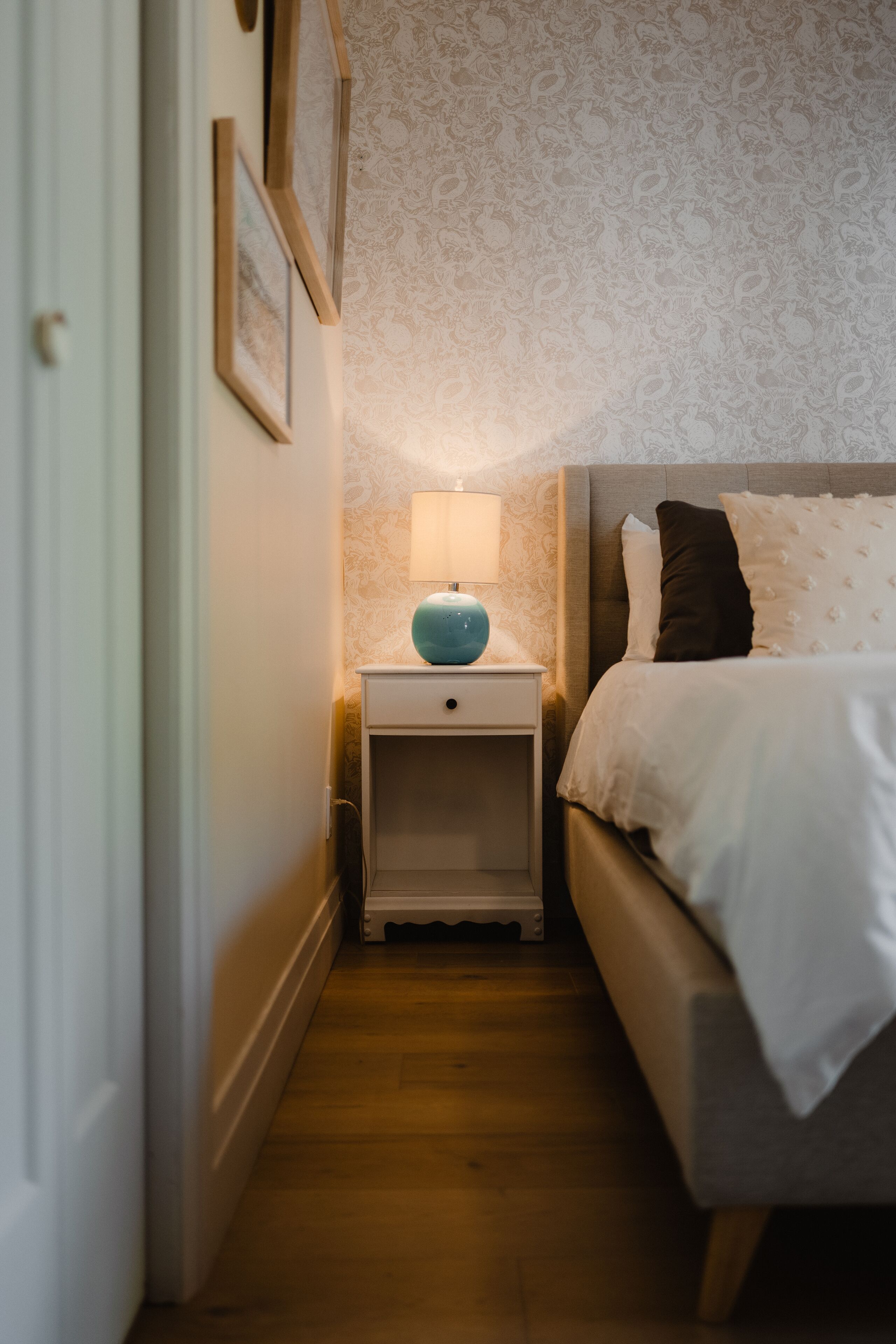 Un pasillo estrecho que conduce a un dormitorio con una cama, una mesita de noche con lámpara y cuadros en la pared.