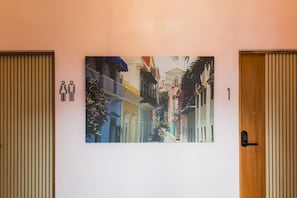A hotel room with a large wall-mounted picture of a colorful street scene, a wooden door with a lock, and a wall-mounted light switch.