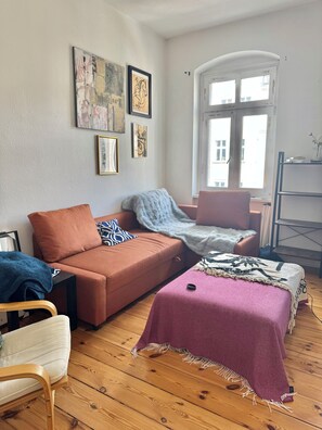 Een knus ogende kamer met een bank, een klein tafeltje, een stoel, een bed met dekbed en kunst aan de wanden.