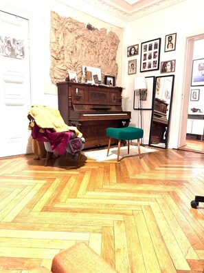A room with a wooden herringbone floor, a piano, a green ottoman, and a wall with framed pictures. - Bright gorgeous Apt, heart of Berlin Mitte (126m2/Three spacious rooms+balcony) (Berlin)
