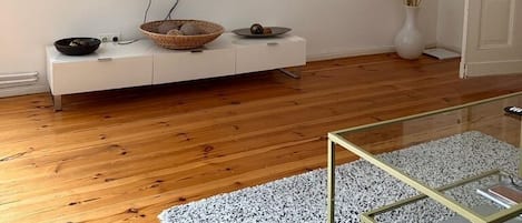 En moderne stue med et fladskærms-tv på væggen, et hvidt tv-møbel med dekorative genstande, et glas sofabord og en grå sofa.